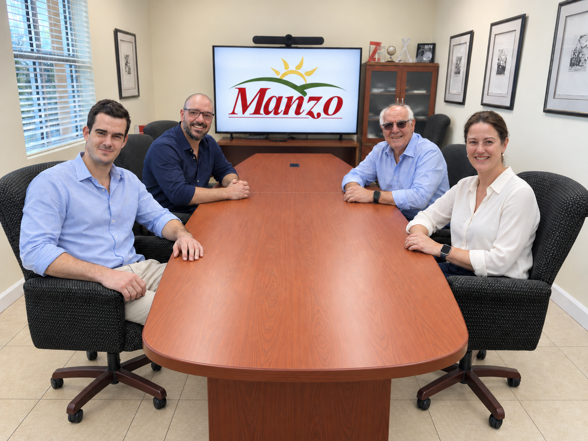 Four people seated around a large wooden conference table in an office, with a screen behind them displaying the Manzo logo.
